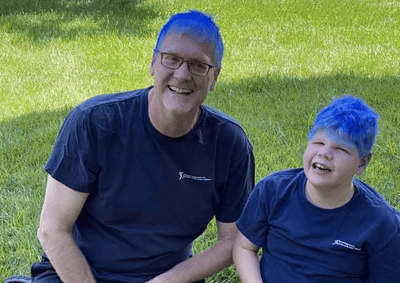 Steve with blue hair with a kid who also has blue hair, in the backyard kneeling in the grass.